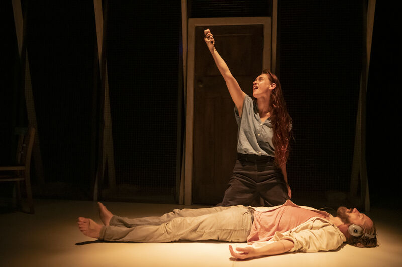 A man lays on the floor still as a woman kneels over him with her left arm raised in the air
