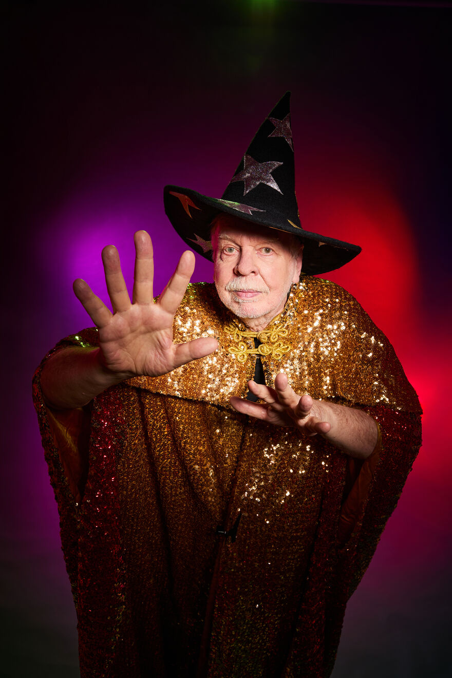Rick Wakeman wearing a gold sequin cape and a wizard hat with his hand outstretched in front of him