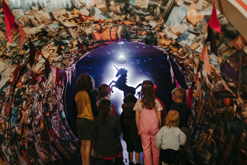  A group of children are gathered together, looking through a tunnel made of colourful, textured fabric. They are looking at a silhouette of a unicorn in front of a bright light. 