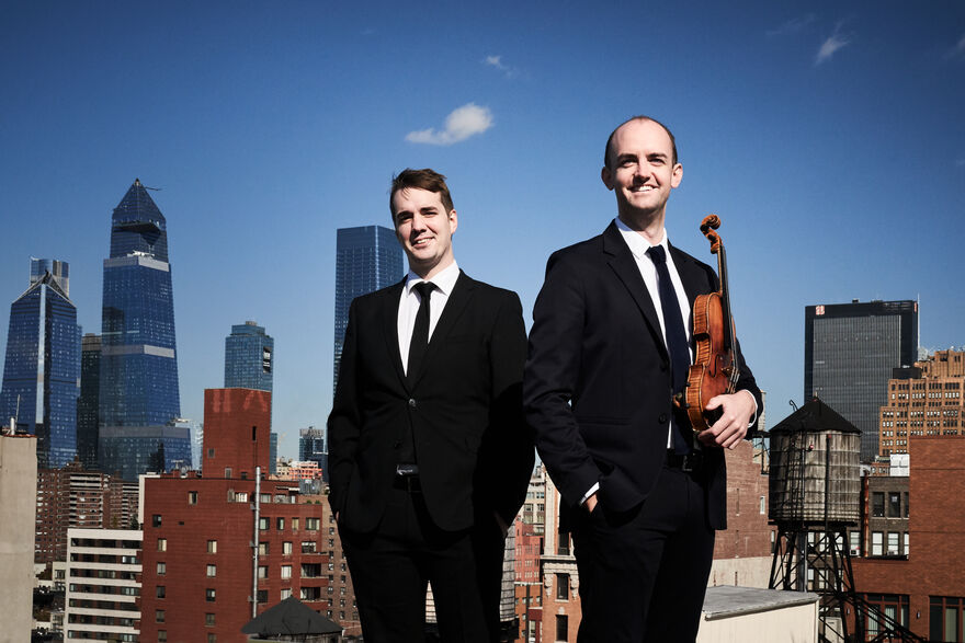 Two men in black suits look directly forwards and smile. The man to the right holds a violin