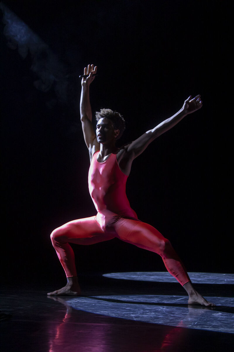 Liam Francis wearing a red unitard in a deep plie in second position with his arms extended above his head