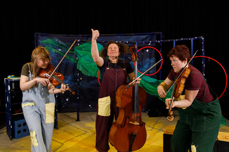 Three people wearing patchwork dungarees playing string instruments enthusiastically