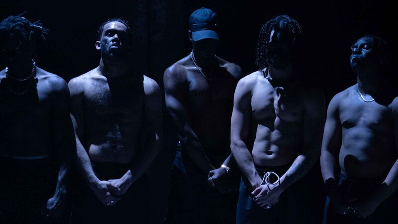 Five intimidating topless men stand on a stage lit by a white light which casts shadows across their chests.