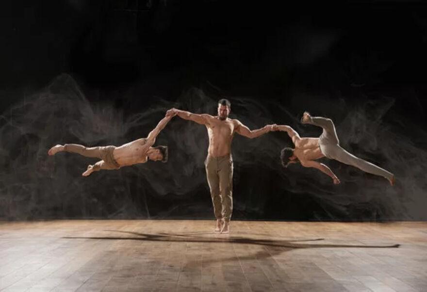 A topless man is holding two other topless men, one in each arm. These men are in the air, doing gymnastics.