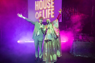 Two performers, one in a suit and one in a long gold dress, sing into microphones. A backdrop behind them spells house of life in large yellow letters