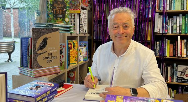 Frank Cottrell-Boyce sat at a desk, surrounded by books, signing a book while smiling