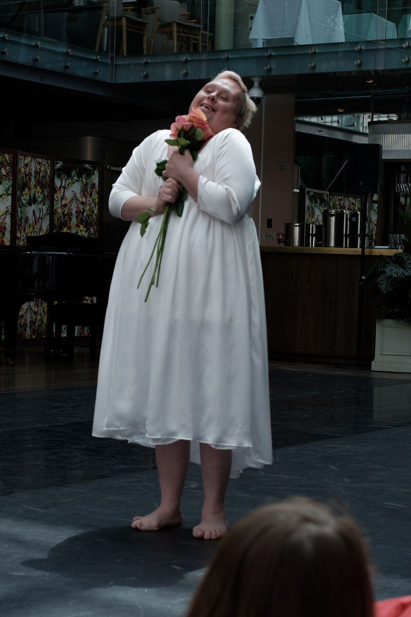 A white performer with Downs Syndrome wears a long white dress and clutches a bunch of roses to their chest