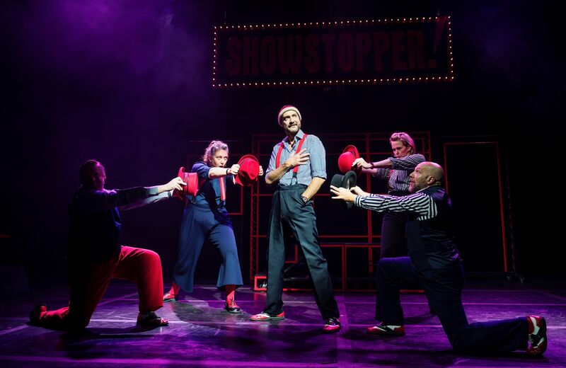 A man in a shirt, trousers and red braces stands centrally, his right hand on his heart, his left in his pocket. He is lit with a spotlight. Around him are a group of 4 people, all gesturing with their hands towards him
