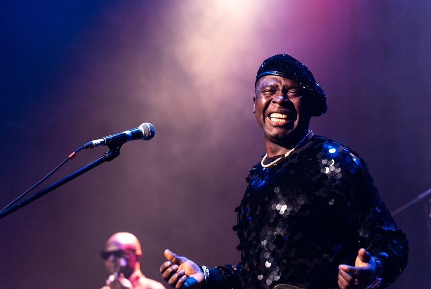 A person singing on stage into a microphone. They are wearing a sequin top and beret