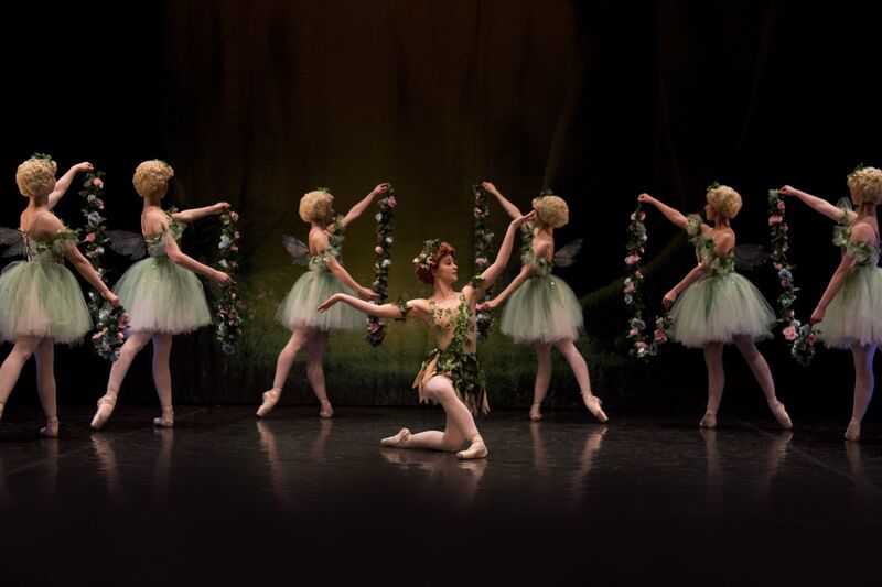 A row of ballerinas in dresses stand en pointe holding up flower garlands. In the middle of them a male ballerina kneels with his arms outstretched