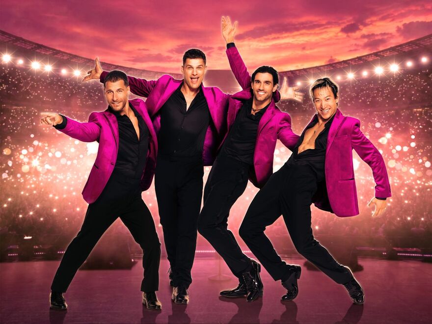 Four men in matching magenta jackets and black outfits pose energetically on a concert stage, with a glowing stadium crowd and vivid pink-orange sunset in the background.