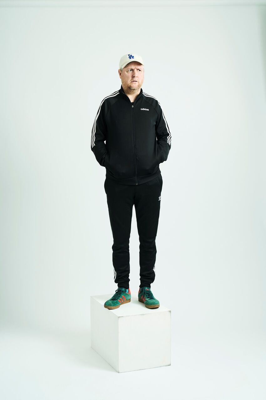 Tim Key stands on a white box against a white backdrop, with his hands in his tracksuit pockets, looking to the side with a blank/confused face