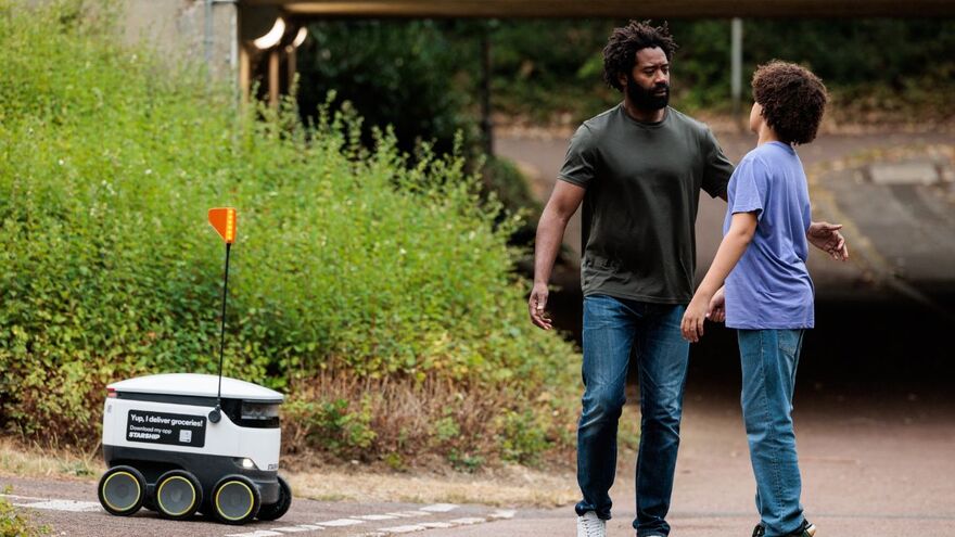 A older man and a younger man are stood opposite each other as though they are about to hug. Next to them is an automated delivery robot