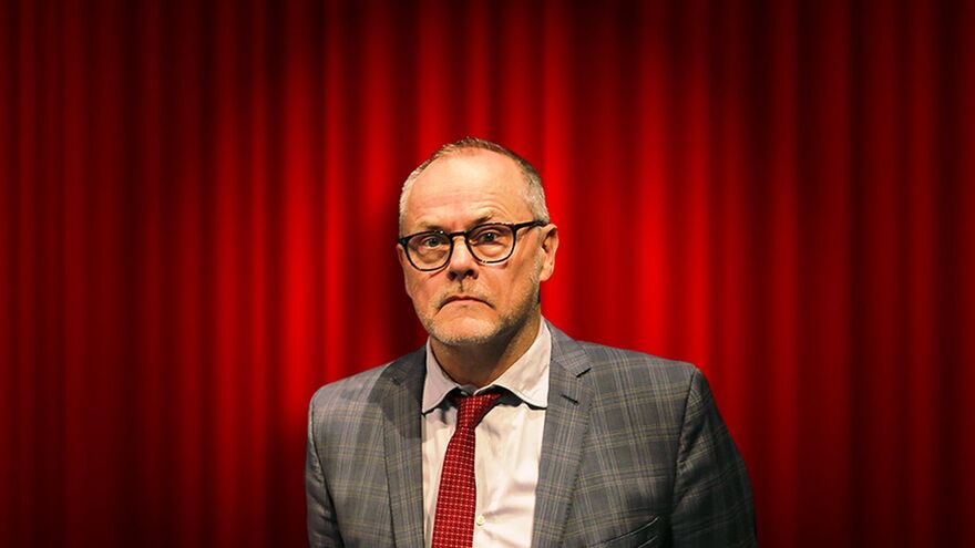 Jack Dee wearing a grey suit and red tie, standing in front of a red theatre curtain