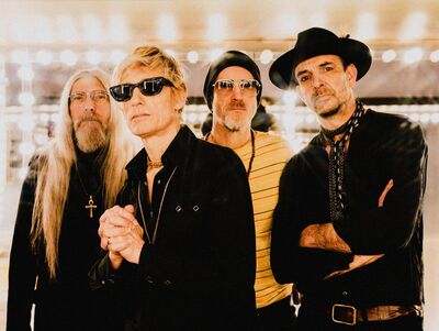 Four people pose together in a warmly lit room, wearing dark outfits, sunglasses, and distinctive accessories. One has long silver hair and a beard, another has short blond hair and clasps his hands, a third wears a yellow striped shirt and black beanie, and the fourth wears a wide-brimmed black hat and patterned scarf.