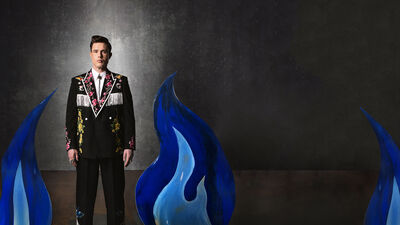Ed Gamble wearing a flowery blazer and matching trousers standing in a dark grey room with cardboard cut outs of blue fire surrounding him