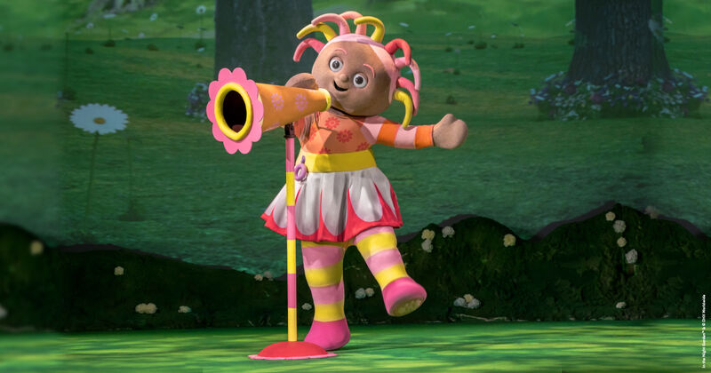 A costumed figure wearing a multi-coloured dress and stripey tights calles into an orange megaphone connected to a stand