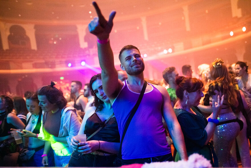 A man in a vest at a club night, his arm is raised, pointing in the air