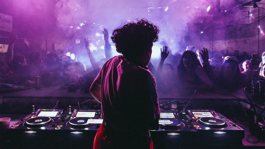 A silhoutte of the back of Annie Mac, standing at some DJ decks wearing a red and pink outfit. In front of her is an audience of clubbers, some with their hands in the air.
