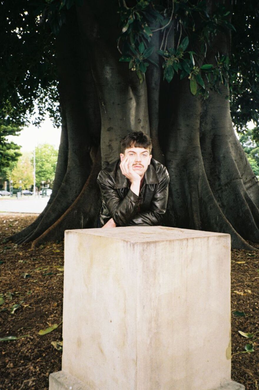 Ed Night leaning on his hand against a concrete cube. He is standing in front of a big tree