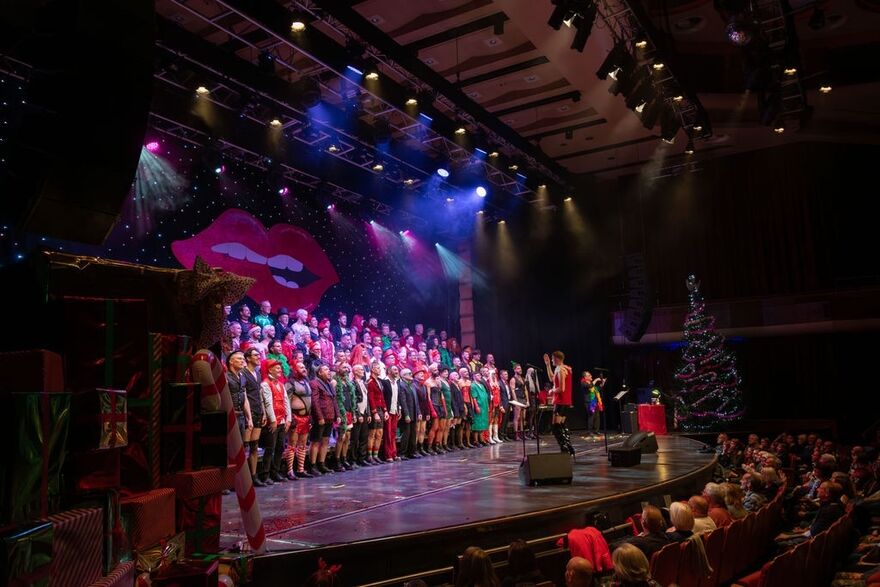 Large choir standing on a stage wearing various christmas outfits. They are standing in front of a big pair of lips.