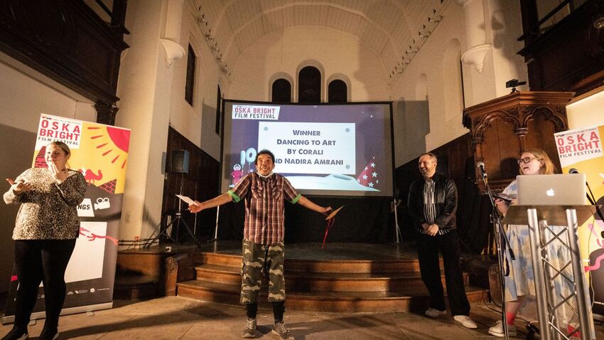 4 people stand in front of a screen. One person is behind a laptop, another is interpreting using British Sign Language and the person in the centre is accepting an award