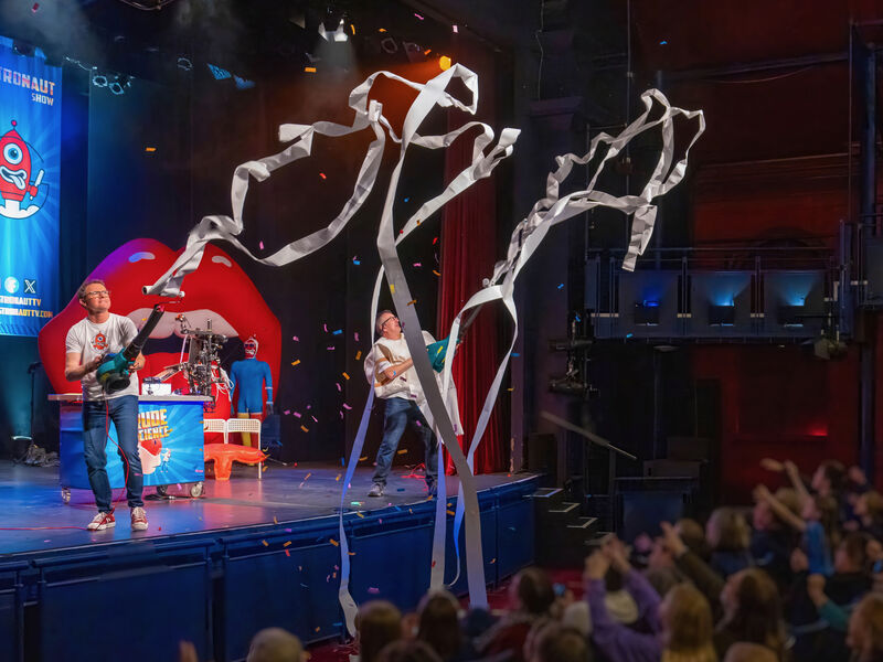 A wide shot shows two men firing confetti canons and rolls of toilet paper into an audience 