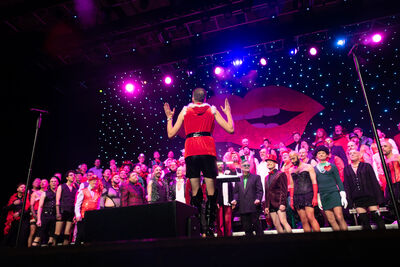 A choir standing on the stage in festive clothing, with a conductor standing with his back to the camera. The stage has purple and blue lights and a cut out of some lips