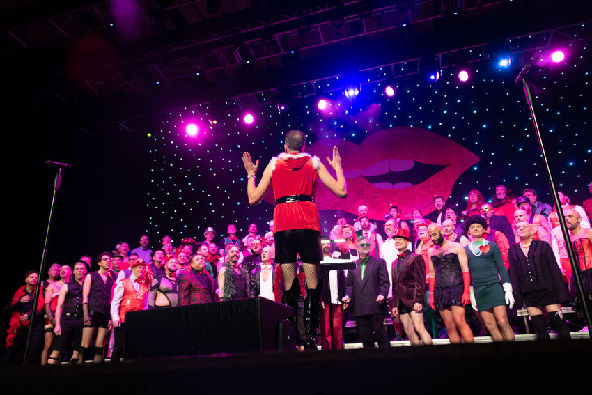 A choir standing on the stage in festive clothing, with a conductor standing with his back to the camera. The stage has purple and blue lights and a cut out of some lips