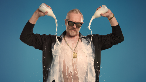 Greg Davies wearing sunglasses, a gold chain and an open jacket over his bare chest. He's pouring two pints of milk down himself
