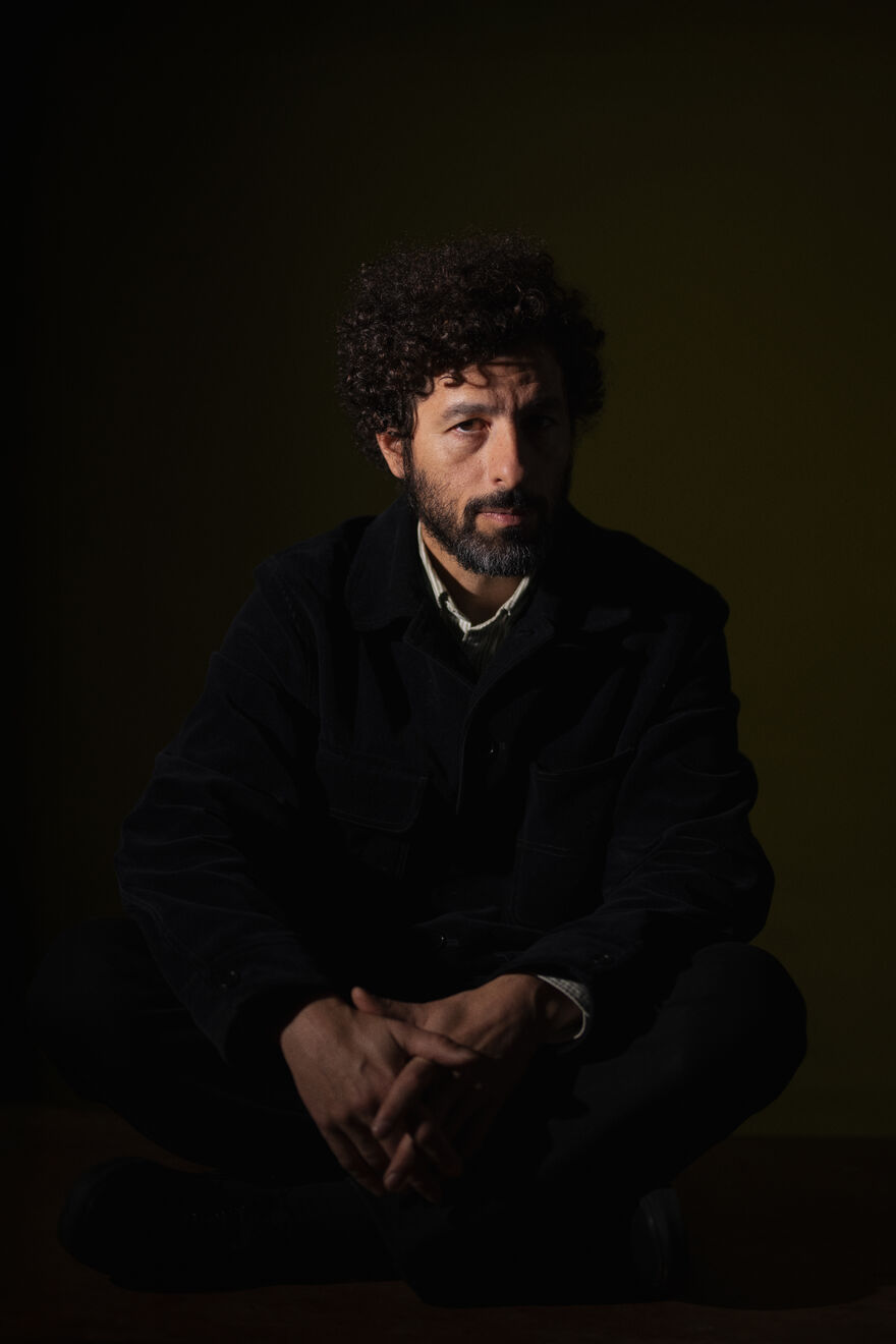 José González wearing a black suit and white shirt, sitting cross legged, half his face is shadowed