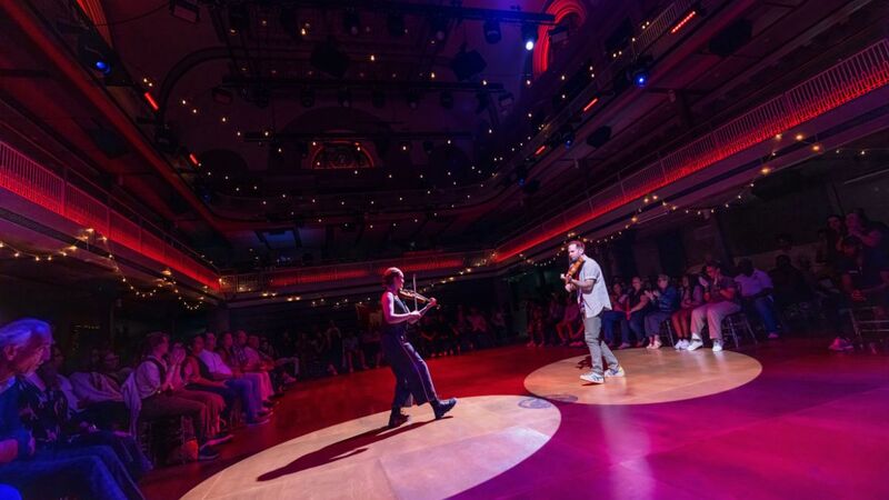 Two musicians face off on stage with string instrument, the audience is sat in the round around them