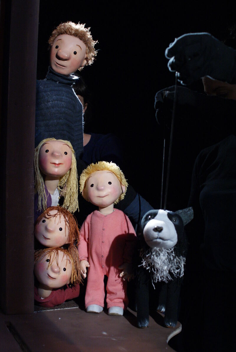 Three wooden puppets of children, their father and a dog poking their heads around the side of a door frame