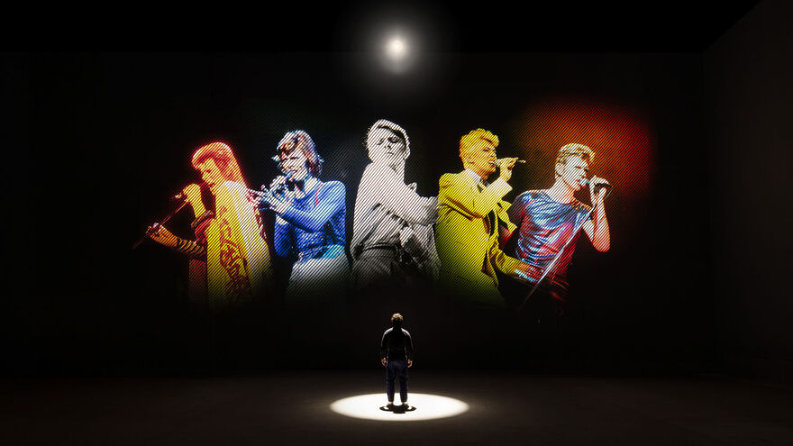 A person stands alone in a dark room beneath a spotlight, facing a large projection of five brightly colored singers performing with microphones.