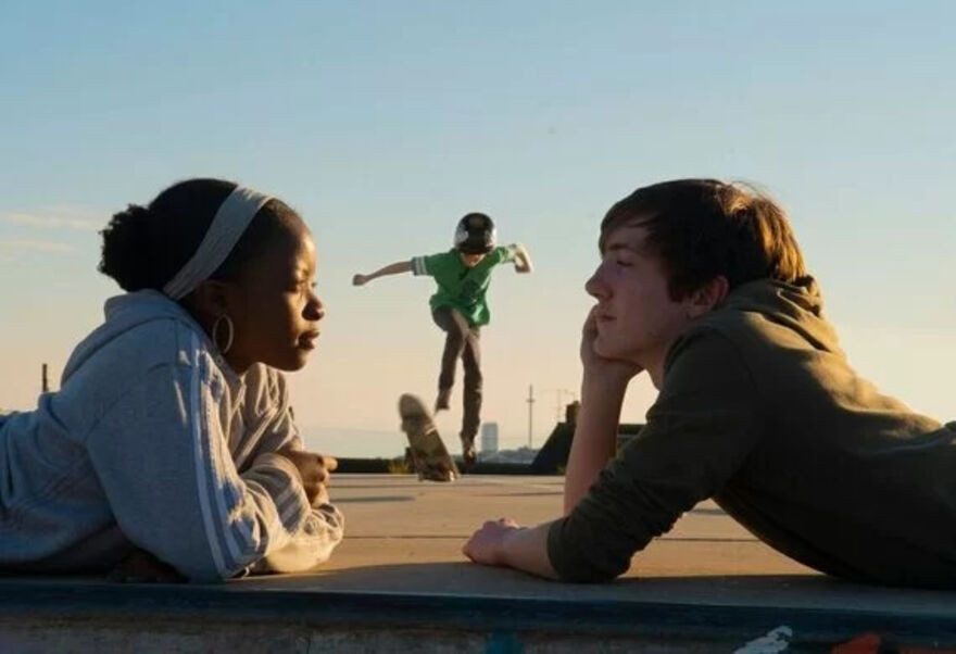 Two young people are looking at eachother whilst laying on their front, behind them a person is doing a skateboard trick