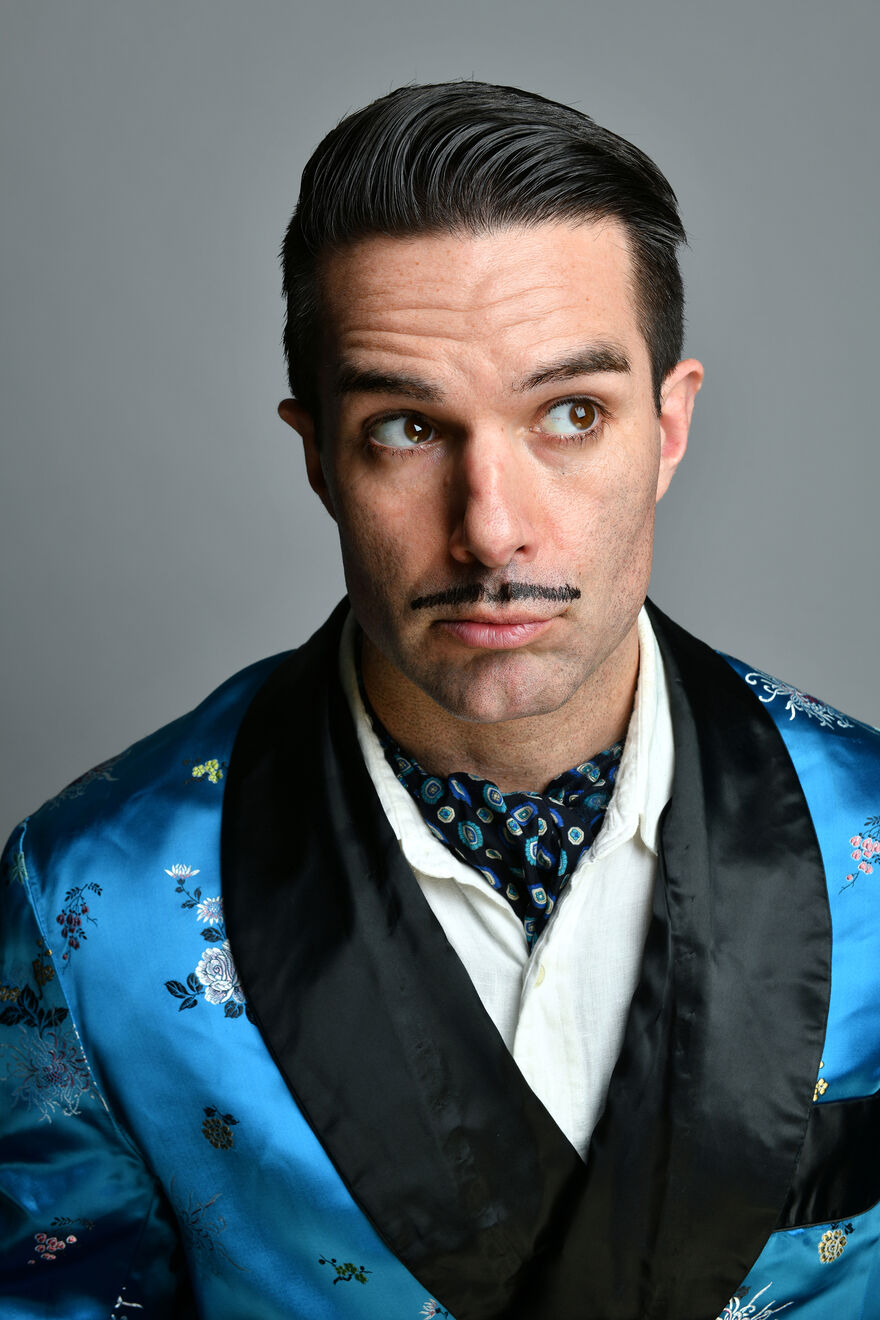 A white man with slicked back black hair and a black pencil moustache. He is wearing a silky blue and black smoking jacket, a white shirt and a cravatte.
