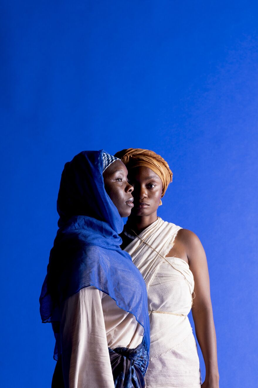 A black woman in a white dress stands, arms by her side, facing forwards. In front of her is another black woman wearing a blue headscarf and white top, she is side-on.