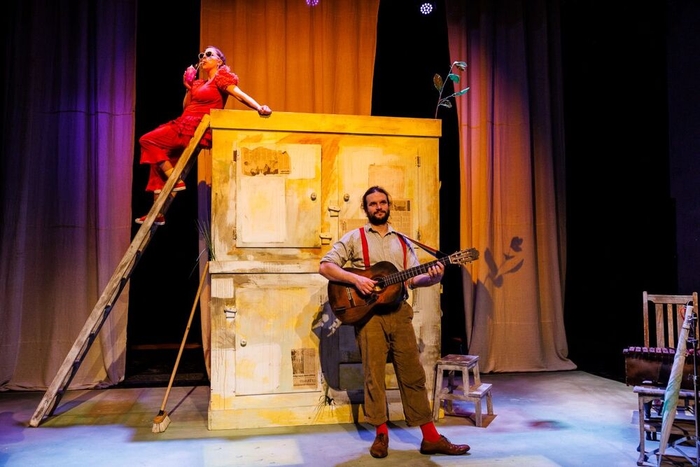 On a stage, there is a big brown box with a woman in red clothes and sunglasses leaning. She is standing on a ladder leaning against the box. In front of the box, there is a man with a bread playing a guitar. The stage is lit in red and purple lights