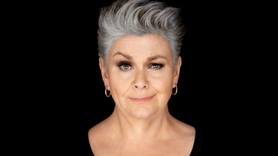 woman white earrings and grey hair looks directly forwards in a dark background