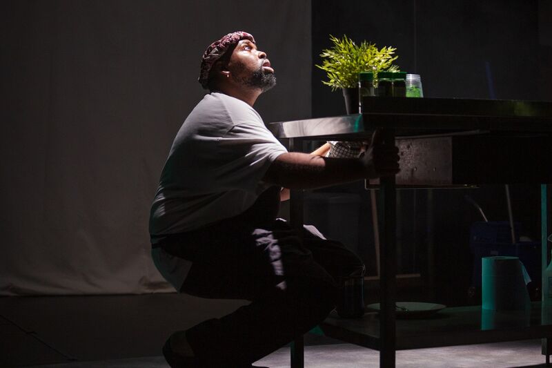 A black man is crouched down behind a desk, holding on to the legs of the desk. There is a lit, green plant on the desk. He looks up toward the light.