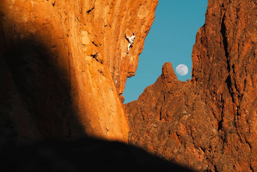 A red mountain with someone climbing up it. It is a dramatic scene with a bright blue sky and the moon peeking through the mountains