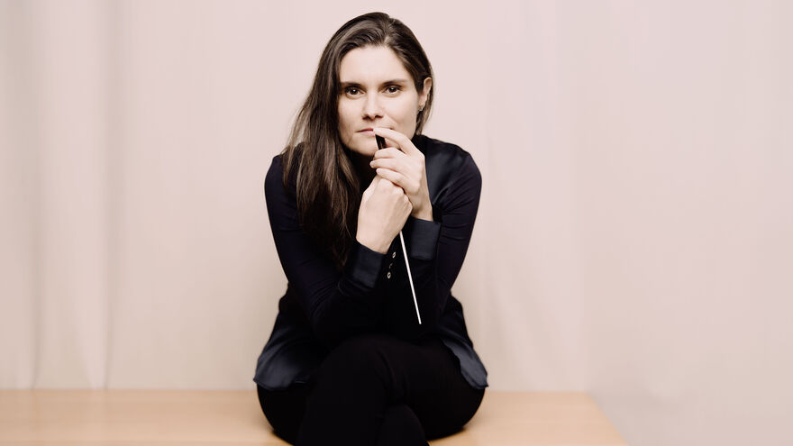 Conductor Delyana Lazarova sits on the edge of a light wooden table against a beige background, wearing a black outfit and holding her baton while looking directly at the camera.