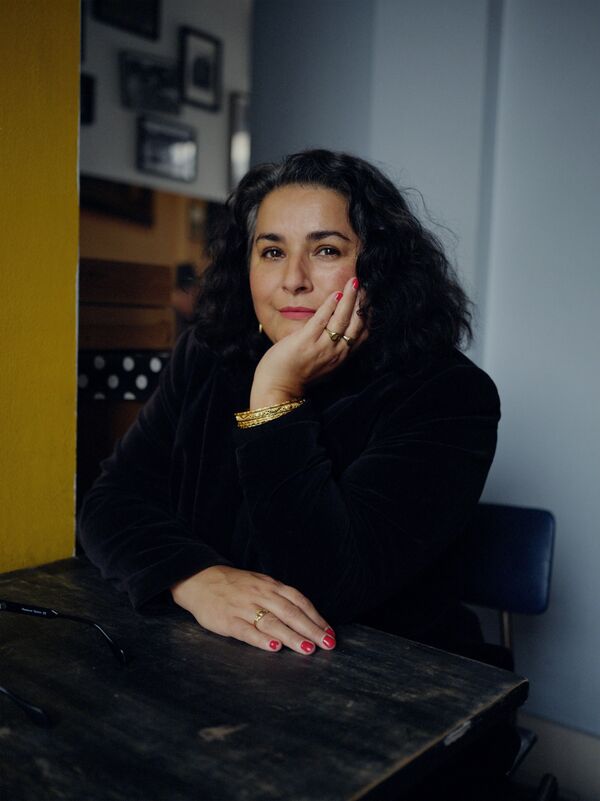 A woman with dark wavy hair sits at a table with one arm resting on the table - her other hand is under her chin