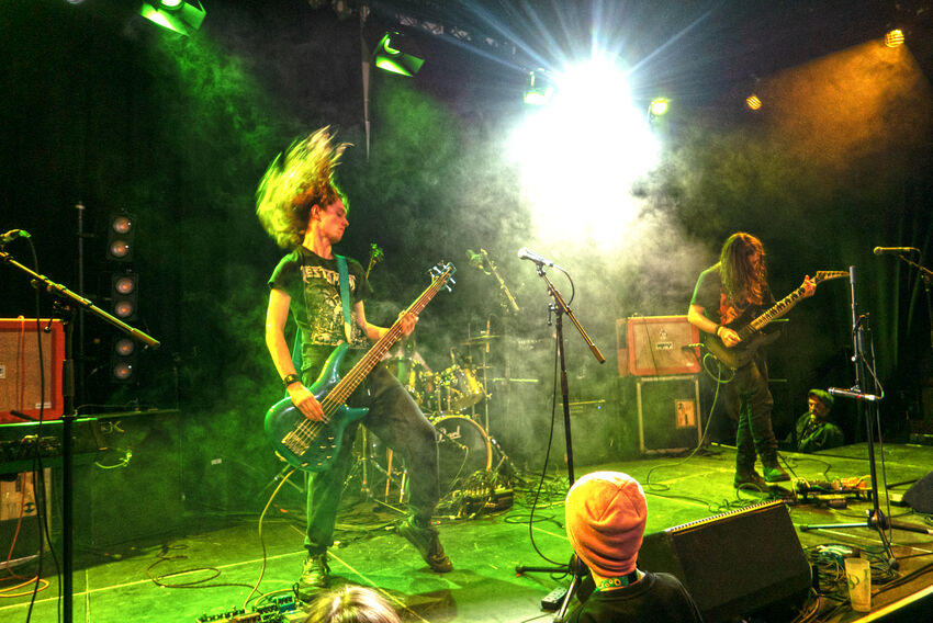 Two people standing on a stage playing a bass and guitar. The stage is lit green and the musican's hair is in the air