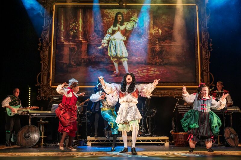 A man dressed as Charles 2nd stands with his arms raised up with two ladies dressed in old-fashioned dresses