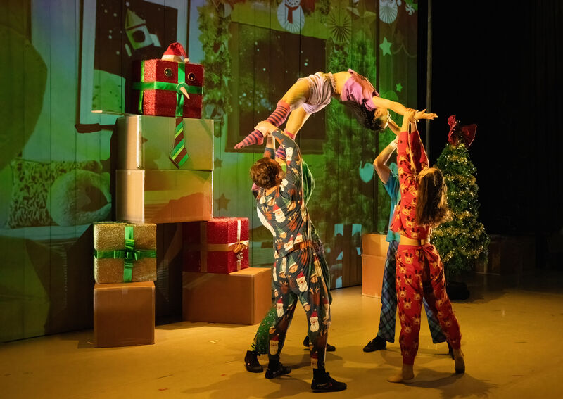 The performers are wearing pjamas holding another performer up in an acrobatic pose with presents in the background