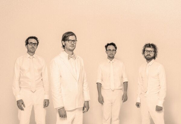 Four men in white suits standing in front of a white wall