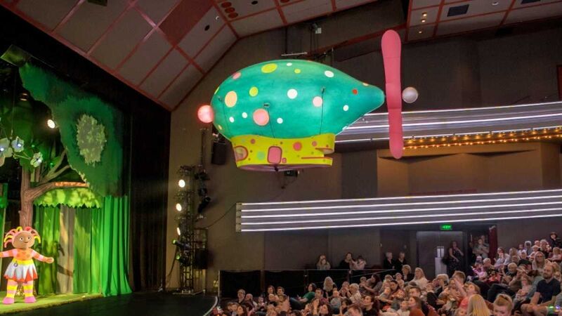A multi-coloured inflatable blimp flies over an auditorium audience towards the stage