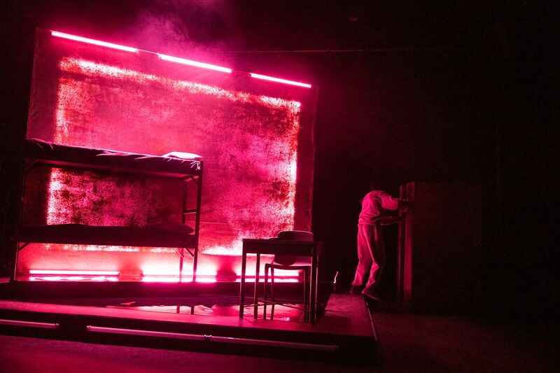 Picture of the set, on one side bunk beds, in the middle a desk and chair, and on the other side Selorm Adonu is standing facing away from the audience. The whole set is bathed in pink light