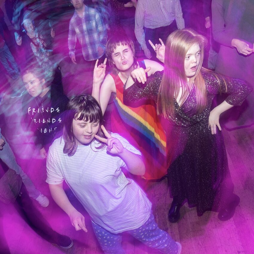 People dancing on a spacious dance floor at a club night. Four people are in the foreground. One is wearing a rainbow dress, one is wearing a 'friends' t-shirt, one has their hand on their hip and one is holding up their hand in a peace sign. The lighting is a blur of blue and purple around them